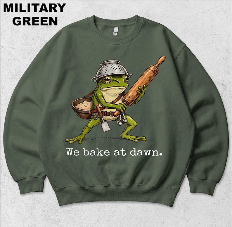 Op de afbeelding: Een militair groen sweatshirt met ronde hals en een afbeelding van een groene kikker die een vergiet als helm draagt, een houten deegroller vasthoudt en een kom en spatel bij zich heeft. De tekst "We bake at dawn." staat in het wit onder de kikker.