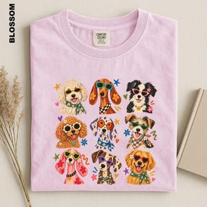 Puede incluir: Una camiseta de cuello redondo color morado claro con un estampado gr&aacute;fico de nueve razas de perros diferentes que llevan gafas de sol coloridas y pa&ntilde;uelos o collares estampados, rodeados de peque&ntilde;os iconos de estrellas, flores y corazones.