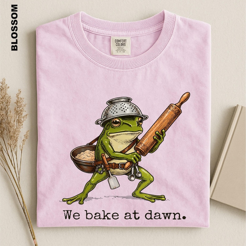 Op de afbeelding: Een lichtroze t-shirt met een afbeelding van een groene kikker die een metalen vergiet als hoed draagt, een houten deegroller vasthoudt en een kleine kom draagt. De tekst "We bake at dawn." staat in het zwart onder de illustratie.