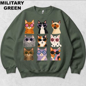 May include: A military green crewneck sweatshirt featuring a graphic print of nine cats arranged in a grid. Each cat wears different colored sunglasses and some have small flowers near their ears. The text MILITARY GREEN appears in the top left.