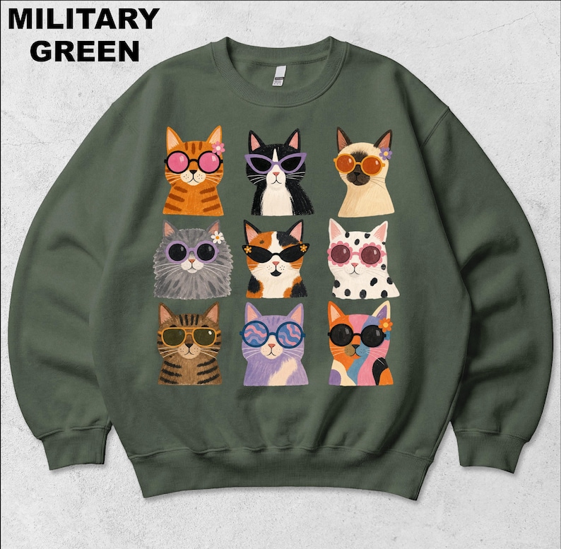 May include: A military green crewneck sweatshirt featuring a graphic print of nine cats arranged in a grid. Each cat wears different colored sunglasses and some have small flowers near their ears. The text MILITARY GREEN appears in the top left.