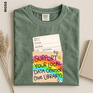 May include: A moss green crew neck t-shirt featuring a graphic of a library checkout card inside a pocket. The design includes a rainbow background with the text "SUPPORT YOUR LOCAL DATA CENTER (THE LIBRARY)" and a small yellow smiley face icon.