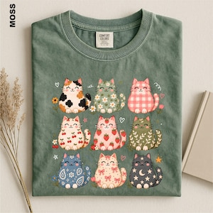 May include: A moss green t-shirt featuring a graphic print of nine stylized cats arranged in a grid. Each cat displays a unique pattern including calico, floral, gingham, cherry, strawberry, leaf, paisley, bow, and moon and star motifs.