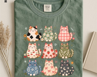 May include: A moss green t-shirt featuring a graphic print of nine stylized cats arranged in a grid. Each cat displays a unique pattern including calico, floral, gingham, cherry, strawberry, leaf, paisley, bow, and moon and star motifs.