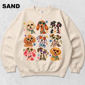 Puede incluir: Una sudadera de cuello redondo color arena con un estampado gr&aacute;fico de nueve razas de perros diferentes. Cada perro lleva gafas de sol coloridas y un pa&ntilde;uelo o collar estampado. La palabra SAND est&aacute; impresa en negro en la esquina superior.