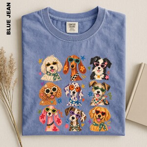 Puede incluir: Una camiseta de color azul vaquero con un estampado gr&aacute;fico de nueve razas de perros diferentes. Cada perro aparece ilustrado con gafas de sol de colores y varios accesorios como pa&ntilde;uelos o flores, rodeado de peque&ntilde;as formas de estrellas y corazones.