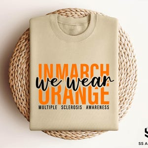 May include: A beige t-shirt with the text "In March We Wear Orange" in orange letters and "Multiple Sclerosis Awareness" in black letters.
