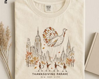 May include: An ivory t-shirt featuring a graphic of a turkey balloon floating above a New York City skyline with a marching band below. The text reads "THANKSGIVING PARADE NEW YORK CITY". The shirt has a Comfort Colors label at the neck.