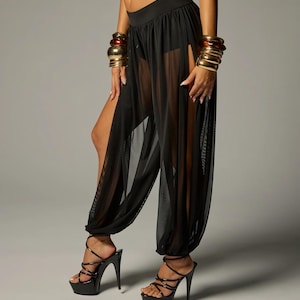 Puede incluir: Pantalones negros transparentes con diseño de cintura alta con capa interior opaca, aberturas laterales anchas que se extienden desde la cadera hasta el dobladillo y puños elásticos fruncidos en los tobillos, con sandalias negras de plataforma.