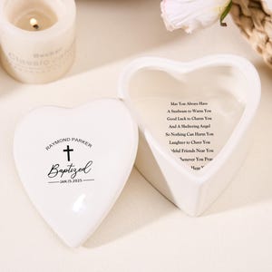 May include: A white heart-shaped keepsake box, open to show an inscription. The lid has the name "Raymond Parker", a cross, and the word "Baptized" with a date. A lit candle and flowers are in the background.