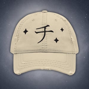 May include: A beige distressed baseball cap featuring a black embroidered Japanese katakana character for chi in the center, flanked by two small four-pointed star icons on the left and one on the right, with a curved brim and stitched eyelets.