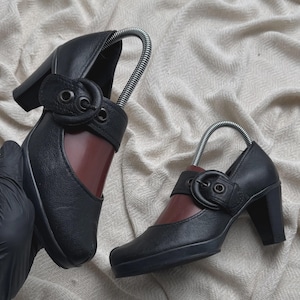 Op de afbeelding: Een paar zwarte leren schoenen met een blokhak en een ronde neus. Elke schoen heeft een brede band over de wreef, vastgezet met een grote, ronde gesp met metalen oogjes. De binnenkant van de schoen heeft een diepe bordeauxrode kleur.