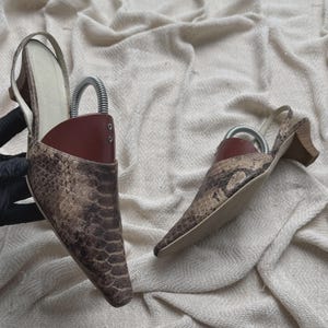 May include: A pair of pointed-toe slingback shoes featuring a brown and tan snakeskin pattern. Each shoe has a low block heel, a light-colored interior lining, and a dark red shoe tree insert with a metal spring mechanism for maintaining shape.