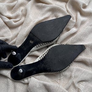 May include: A pair of pointed-toe mules featuring black outsoles and small kitten heels. The upper portion of the footwear displays a white woven mesh pattern with a distinct texture. The soles show signs of wear, and the heels have silver-toned caps.