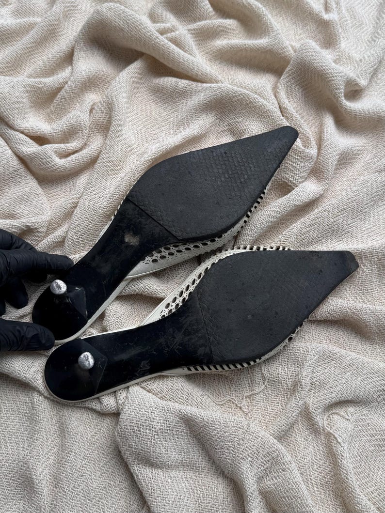 May include: A pair of pointed-toe mules featuring black outsoles and small kitten heels. The upper portion of the footwear displays a white woven mesh pattern with a distinct texture. The soles show signs of wear, and the heels have silver-toned caps.