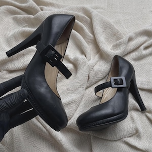 Puede incluir: Un par de zapatos de tacón alto negros con punta redondeada, suela de plataforma y una correa en el empeine asegurada con una hebilla cuadrada. Los zapatos tienen un acabado liso y un tacón de aguja alto con un diseño clásico.