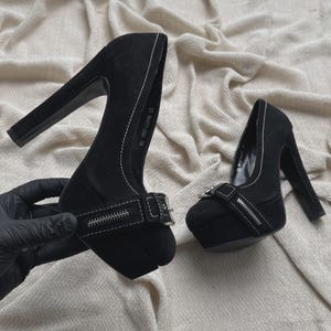 Pode incluir: Um par de sapatos de salto alto com plataforma na cor preta, com salto bloco grosso, bico arredondado e costuras brancas em contraste. Cada sapato inclui uma tira decorativa com fivela de metal prateado e um pequeno detalhe de zíper.