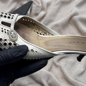May include: A white leather mule shoe featuring a laser-cut perforated pattern across the upper, a circular decorative accent near the opening, and a tan interior insole with the number 40 printed clearly on the side of the inner heel area.