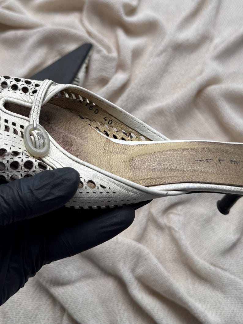 May include: A white leather mule shoe featuring a laser-cut perforated pattern across the upper, a circular decorative accent near the opening, and a tan interior insole with the number 40 printed clearly on the side of the inner heel area.