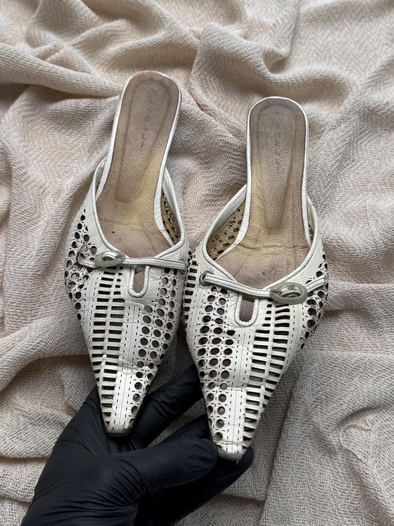 May include: A pair of white pointed-toe mules featuring a laser-cut geometric pattern across the upper. Each shoe includes a decorative silver-tone metal buckle detail at the vamp and a gold-toned interior footbed with a visible size 40 marking.