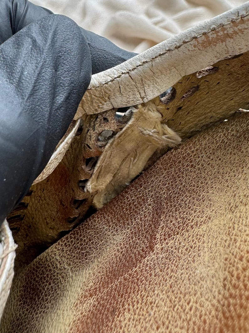 May include: A close-up view of a tan leather item featuring a white leather trim with visible cracking and wear. The edge shows a decorative perforated pattern with brown interior detailing and frayed fabric stitching visible along the seam.