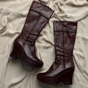 Peut inclure: Une paire de bottes compensées marron foncé, arrivant au genou. Les bottes ont une semelle plateforme et une garniture décorative sur le dessus et sur le devant. Elles sont en matière lisse semblable au cuir.