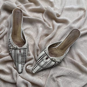 May include: A pair of off-white pointed-toe mules featuring a laser-cut geometric pattern across the vamp. Each shoe includes a decorative horizontal strap with a small oval buckle detail at the center and a tan-colored interior footbed.