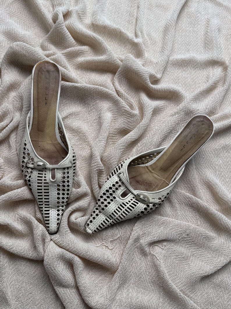May include: A pair of off-white pointed-toe mules featuring a laser-cut geometric pattern across the vamp. Each shoe includes a decorative horizontal strap with a small oval buckle detail at the center and a tan-colored interior footbed.