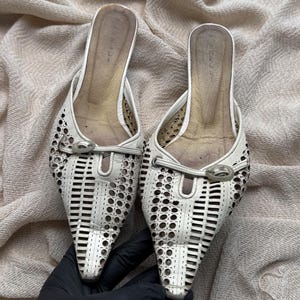 May include: A pair of white pointed-toe mules featuring a laser-cut geometric pattern across the upper. Each shoe includes a decorative silver-tone metal buckle detail at the vamp and a gold-toned interior footbed with a visible size 40 marking.