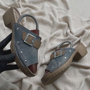 May include: A pair of open-toe mules featuring a blue denim-like upper with silver-tone metal studs and a brown strap with a silver buckle. The footwear has a brown block heel and a textured sole, with a metal shoe tree insert visible inside.
