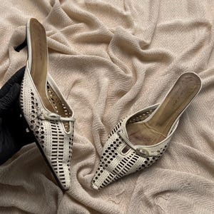 May include: A pair of off-white pointed-toe mules featuring intricate laser-cut geometric patterns across the upper. Each shoe includes a small decorative buckle detail at the vamp and a slim kitten heel. The interior insoles are tan.