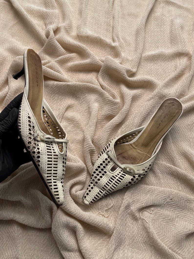 May include: A pair of off-white pointed-toe mules featuring intricate laser-cut geometric patterns across the upper. Each shoe includes a small decorative buckle detail at the vamp and a slim kitten heel. The interior insoles are tan.