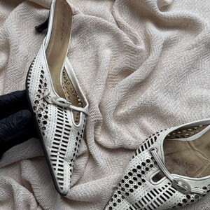 May include: A pair of white pointed-toe mules featuring a laser-cut geometric pattern across the upper, a small kitten heel, and a decorative oval buckle detail on the vamp. The interior footbed is tan, and the shoes have a structured design.