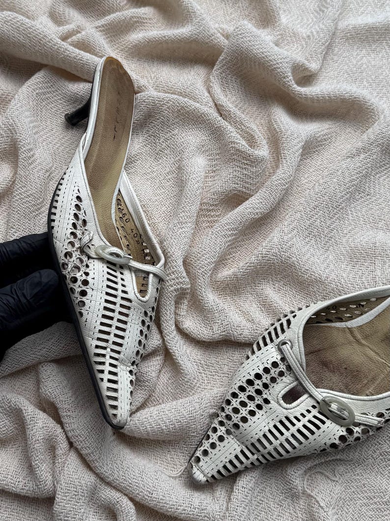May include: A pair of white pointed-toe mules featuring a laser-cut geometric pattern across the upper, a small kitten heel, and a decorative oval buckle detail on the vamp. The interior footbed is tan, and the shoes have a structured design.
