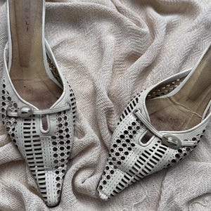 May include: A pair of off-white pointed-toe mules featuring intricate laser-cut circular and rectangular patterns across the vamp. Each shoe includes a decorative oval buckle detail at the center and a tan-colored interior footbed.