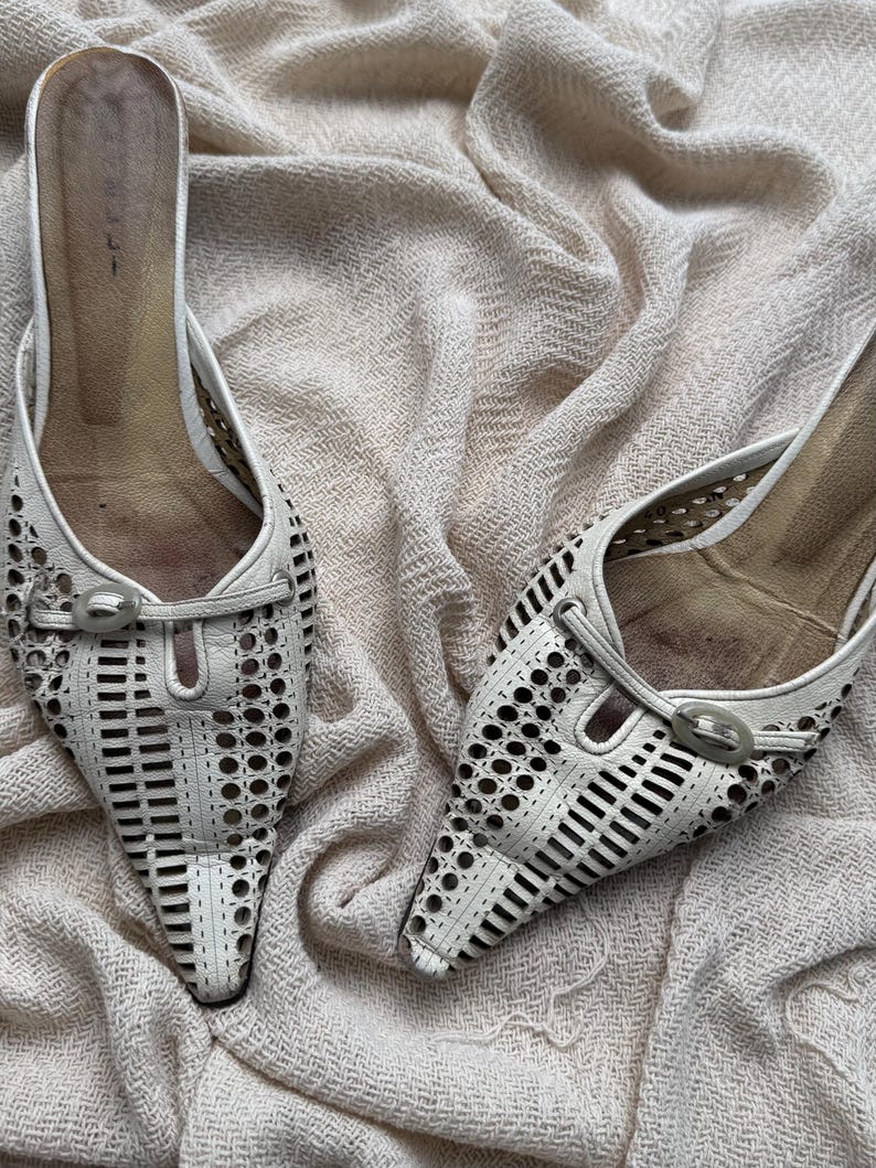 May include: A pair of off-white pointed-toe mules featuring intricate laser-cut circular and rectangular patterns across the vamp. Each shoe includes a decorative oval buckle detail at the center and a tan-colored interior footbed.