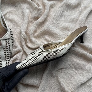 May include: A white leather mule with a pointed toe and a kitten heel. The upper features a laser-cut geometric pattern throughout. The interior sole is tan and displays the size 40 printed in black. The shoe has a slim black heel base.