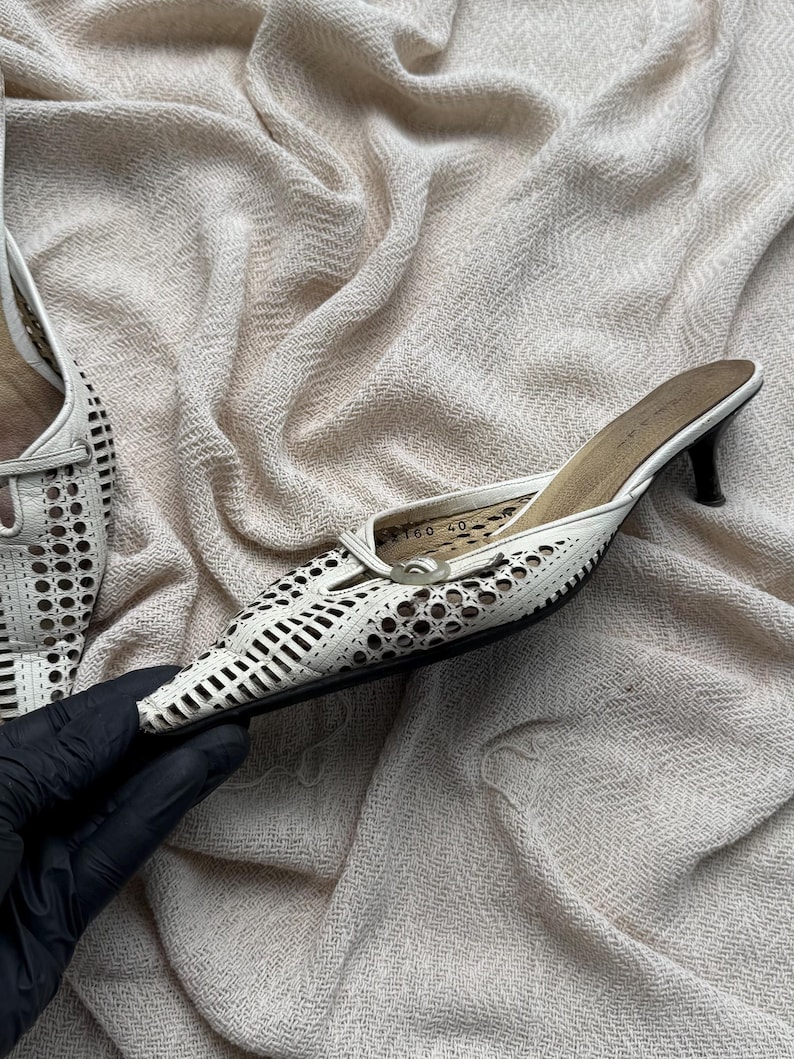 May include: A white leather mule with a pointed toe and a kitten heel. The upper features a laser-cut geometric pattern throughout. The interior sole is tan and displays the size 40 printed in black. The shoe has a slim black heel base.
