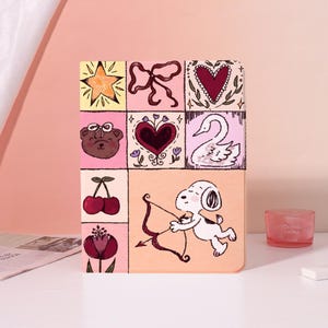 May include: A rectangular tablet case featuring a grid of illustrations including a yellow star, a brown bear with a bow, red cherries, a dark red flower, a maroon heart, a white swan, and a white dog holding a bow and arrow on a peach background.