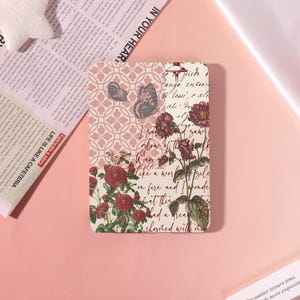 May include: A rectangular e-reader case featuring a collage design with a pink geometric pattern, two butterflies, red roses, and cursive script text. The background includes a cream-colored section with handwritten-style black lettering.