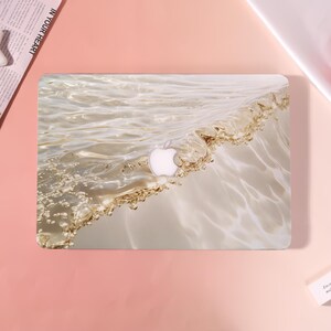 May include: A laptop case with a wave design in beige and gold tones. The Apple logo is visible. The case has a glossy finish. The background is pink. The case is designed to fit a laptop.