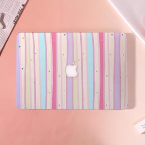 May include: A laptop skin featuring a pastel vertical stripe design in pink, purple, blue, and cream colors. The Apple logo is visible in the center. Small, colorful dots are scattered across the surface, adding a playful touch to the overall aesthetic.