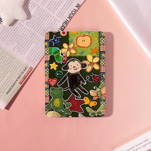 May include: A rectangular green notebook cover with a whimsical design featuring a monkey, flowers, stars, and a frog. The cover has the text "I LOVE YOU!" and "I LOVE". The background is a mix of green and orange.