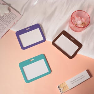 May include: Three rectangular ID badge holders in purple, teal, and brown, each with a white insert. A small pink glass with clear crystals and a matchbox with text that reads "Encounter leisure time. make life more exquisite" are also present.