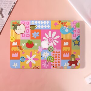 May include: A laptop skin featuring a colourful patchwork design with various icons including an apple, a blue fish, a red tomato, a green frog on a mushroom, flowers, stars, buttons, and a central cutout for an Apple logo on a rectangular surface.