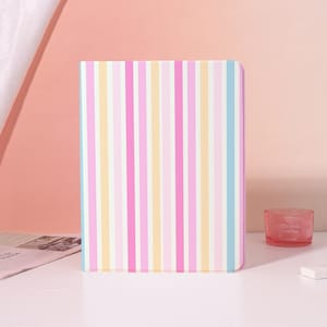 May include: A pastel-colored striped notebook with vertical stripes in pink, yellow, light blue, and white. The notebook is rectangular with rounded corners. A small, round, pink glass jar with the text "CRYSTAL AROMA" is visible in the background.