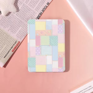 Peut inclure: Un étui rectangulaire avec un motif patchwork aux couleurs pastel, notamment des carrés roses, jaunes, bleus et verts. Certains carrés présentent des motifs à pois ou à carreaux. De petits boutons ronds sont fixés. Le texte "LIFE IS LIKE A CAFETERIA" est visible.