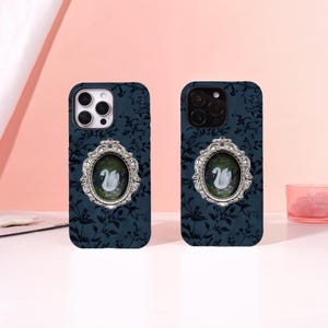May include: Two dark blue smartphone cases featuring a repeating black floral pattern. Each case displays a central oval-shaped silver frame with ornate detailing, enclosing a small illustration of a white swan swimming on dark green water.