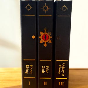 May include: Three hardcover books with black covers and gold lettering. The titles are "Horus Rising," "False Gods," and "Galaxy in Flames." Each book has a gold symbol and a bookmark.