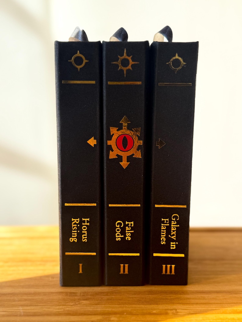 May include: Three hardcover books with black covers and gold lettering. The titles are "Horus Rising," "False Gods," and "Galaxy in Flames." Each book has a gold symbol and a bookmark.